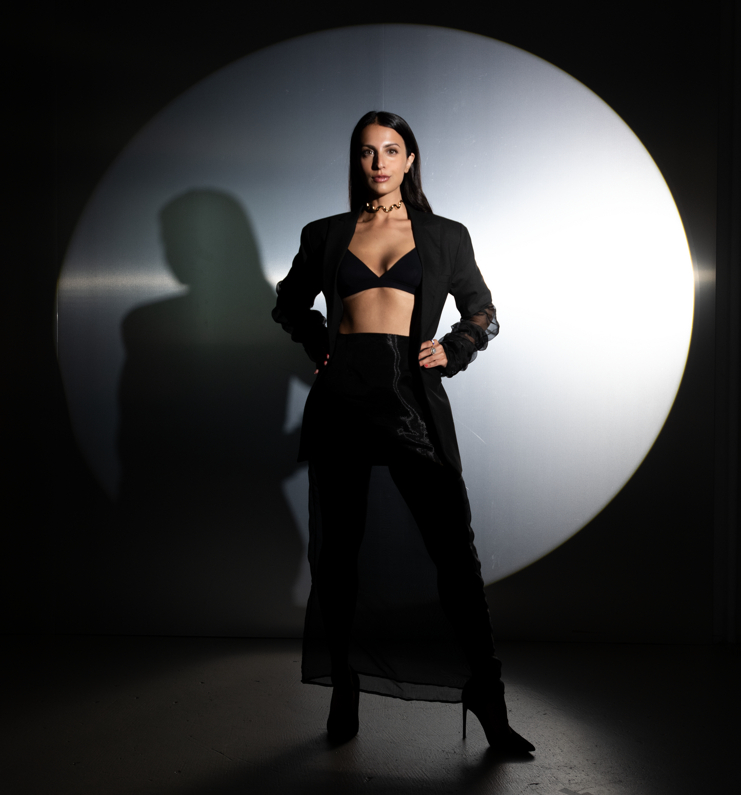 Woman wearing a black bralette, long black blazer, and fitted skirt with a layered mesh bottom, standing confidently in front of a large circular spotlight.
